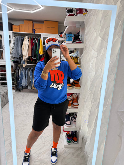 Person taking a mirror selfie wearing a blue sweatshirt with red and white text, black shorts, and orange socks in a room with shelves and clothing.